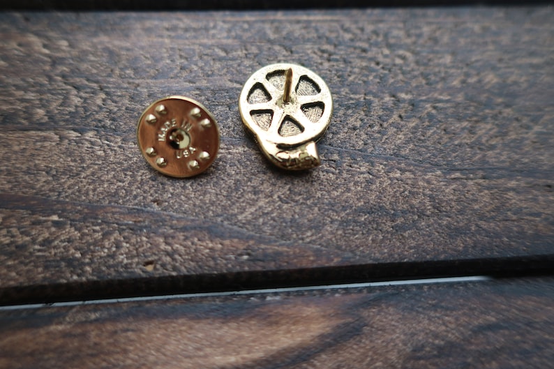May include: Gold-toned metal film reel pin with a pin back. The back of the pin has the words "Made in USA" on it.