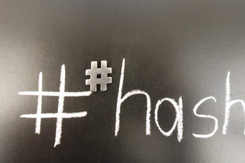 May include: A white hashtag symbol drawn on a black chalkboard with a silver metal hashtag charm placed on top of the drawn symbol. The word "hash" is written in white chalk below the symbol.