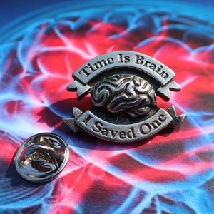 K&ouml;nnte beinhalten: Silberfarbener Anstecker mit einem Gehirndesign und dem Text "Time Is Brain I Saved One". Der Anstecker befindet sich auf einem blauen und roten Hintergrund.