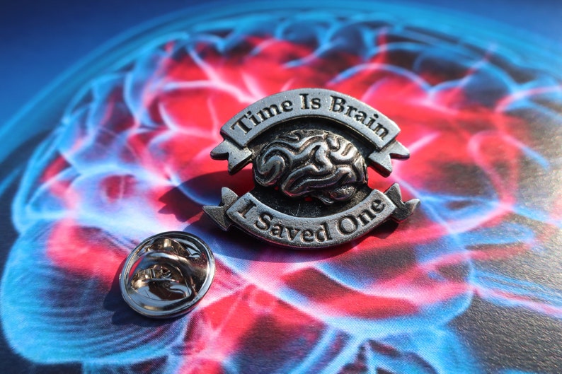 K&ouml;nnte beinhalten: Silberfarbener Anstecker mit einem Gehirndesign und dem Text "Time Is Brain I Saved One". Der Anstecker befindet sich auf einem blauen und roten Hintergrund.