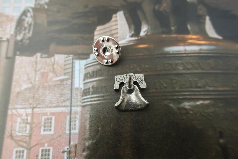 Puede incluir: Pin de plata de la Campana de la Libertad con un reverso que dice "Clif USA". El pin se muestra sobre un fondo de una foto de la Campana de la Libertad.