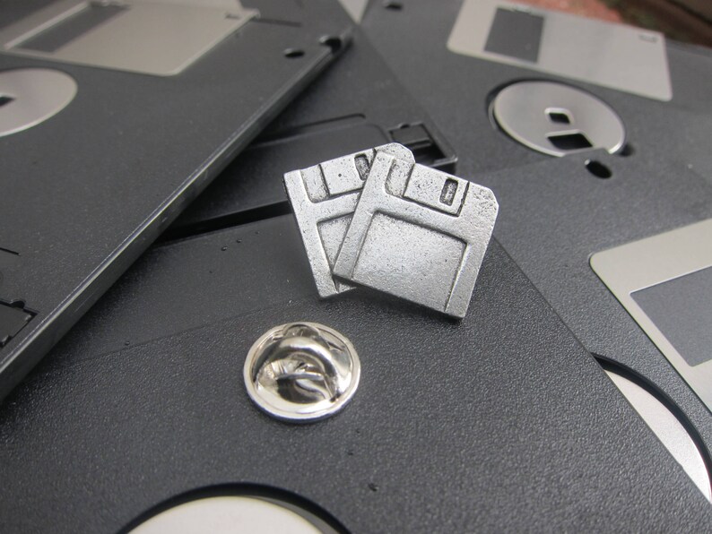 May include: Two silver-colored floppy disk pins and a silver lapel pin rest on a background of black floppy disks. The pins are detailed with the classic floppy disk design, and the lapel pin has a rounded shape.