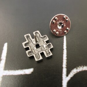May include: Silver-toned metal pin shaped like a hashtag symbol. The back of the pin has a circular metal backing with the words "Made in USA".