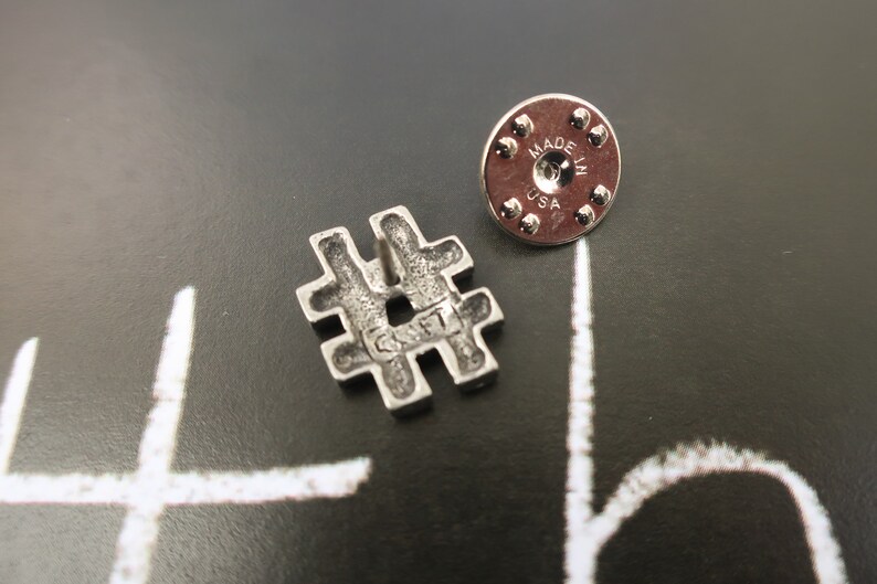 May include: Silver-toned metal pin shaped like a hashtag symbol. The back of the pin has a circular metal backing with the words "Made in USA".