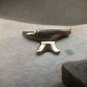 Peut inclure: Un petit charme en m&eacute;tal argent&eacute; en forme d'enclume. L'enclume est un outil traditionnel de forgeron.