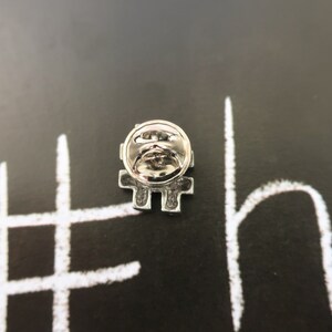 May include: Silver pin with a hashtag design. The pin is on a black background with white chalk writing that says "#" and "ho".