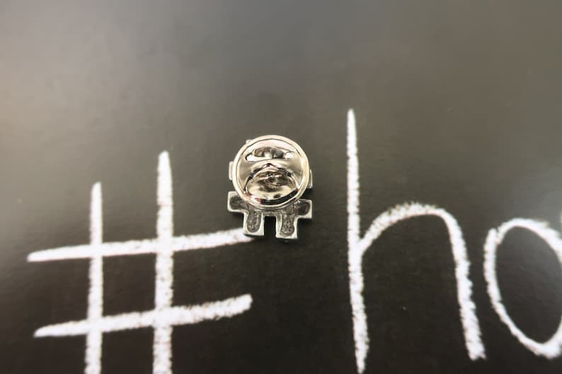 May include: Silver pin with a hashtag design. The pin is on a black background with white chalk writing that says "#" and "ho".