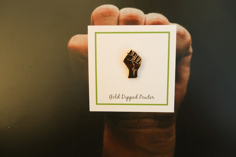 May include: A gold-dipped pewter pin featuring a raised fist. The pin is on a white card with green trim and the text "Gold Dipped Pewter".