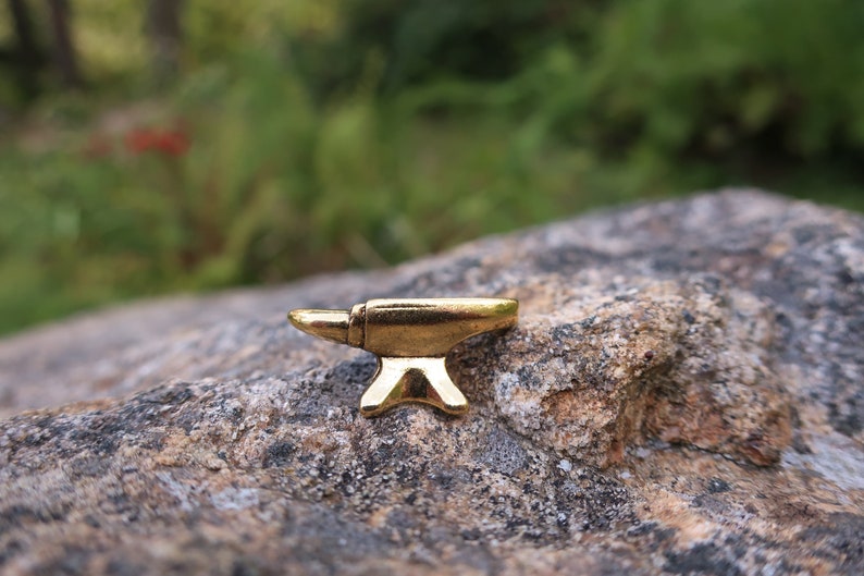 May include: A small gold-colored metal anvil on a gray rock surface.