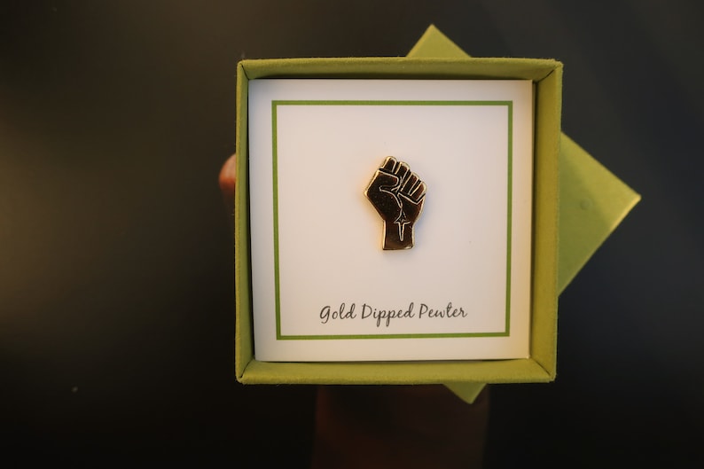 May include: A gold-dipped pewter pin in the shape of a raised fist. The pin is in a green box with a white card that says "Gold Dipped Pewter".