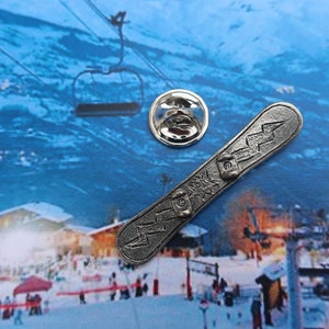Puede incluir: Un pin de metal plateado con forma de tabla de snowboard con un dise&ntilde;o de rayo. El pin est&aacute; sobre un fondo de escena invernal con un remonte y monta&ntilde;as cubiertas de nieve.
