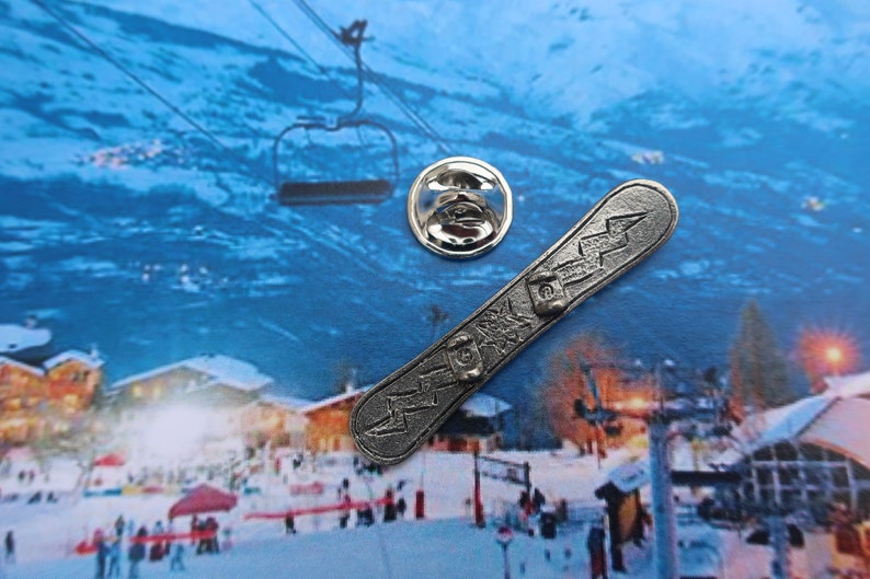Puede incluir: Un pin de metal plateado con forma de tabla de snowboard con un dise&ntilde;o de rayo. El pin est&aacute; sobre un fondo de escena invernal con un remonte y monta&ntilde;as cubiertas de nieve.