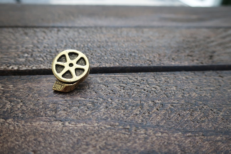 May include: A gold-colored film reel charm on a wooden surface.