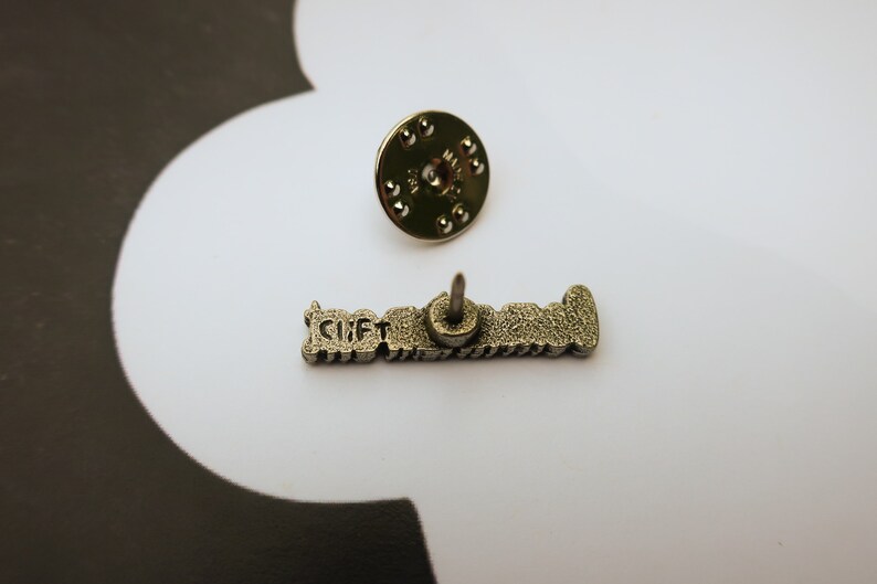 May include: Silver-toned metal pin with the text "GIFT" in raised letters. The pin has a back post and a butterfly clutch.