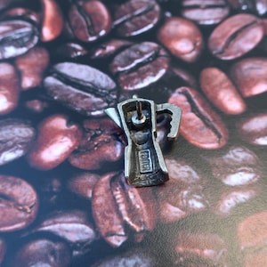 May include: Silver-toned metal charm in the shape of a traditional Italian coffee pot, or Moka pot, with a detailed design. The charm is sitting on a bed of coffee beans.