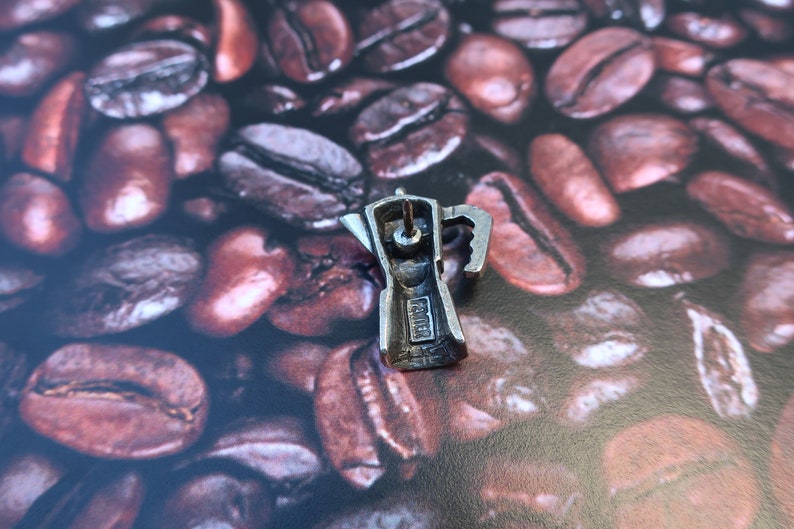 May include: Silver-toned metal charm in the shape of a traditional Italian coffee pot, or Moka pot, with a detailed design. The charm is sitting on a bed of coffee beans.