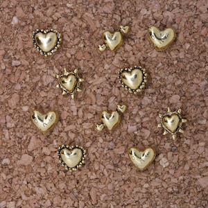 Puede incluir: Un tablero de corcho con diez alfileres de cabeza dorada en forma de corazón. Los alfileres tienen diferentes diseños, incluyendo un sol, un corazón con una flecha y un corazón con un remolino.