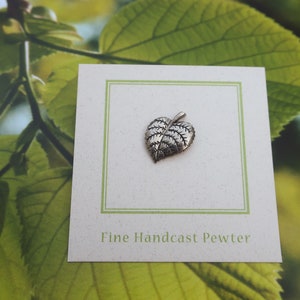 May include: A small, detailed pewter leaf pendant on a white card with a green border. The card is set against a backdrop of green leaves. The text "Fine Handcast Pewter" is printed on the card.