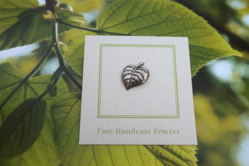 May include: A small, detailed pewter leaf pendant on a white card with a green border. The card is set against a backdrop of green leaves. The text "Fine Handcast Pewter" is printed on the card.