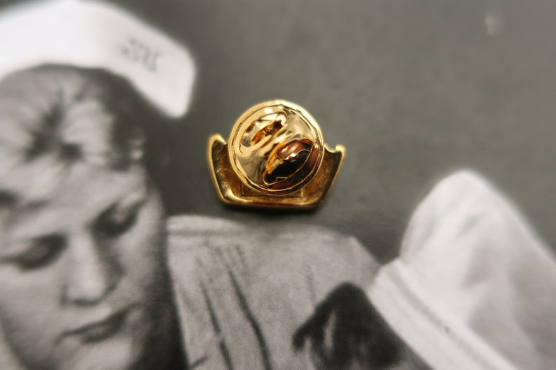 May include: Gold-toned pin with a circular design. The pin has a safety clasp on the back.