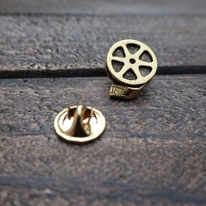 May include: Gold-toned film reel pin with a decorative back. The pin is on a dark brown wooden surface.