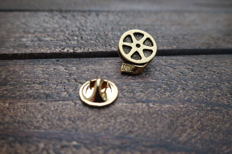 May include: Gold-toned film reel pin with a decorative back. The pin is on a dark brown wooden surface.