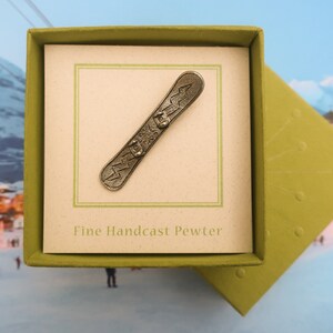 Puede incluir: Un amuleto de snowboard de peltre en una caja de regalo verde. La caja tiene las palabras "Fine Handcast Pewter" impresas en el interior.
