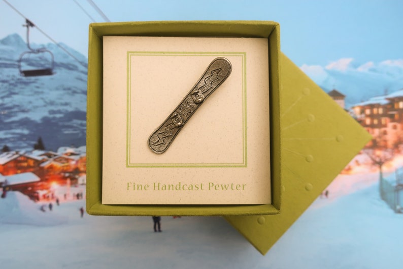 Puede incluir: Un amuleto de snowboard de peltre en una caja de regalo verde. La caja tiene las palabras "Fine Handcast Pewter" impresas en el interior.