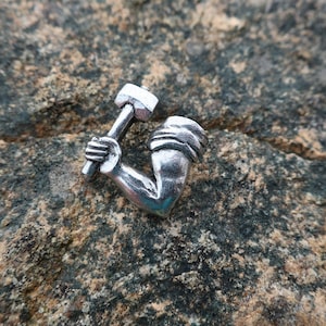 May include: A silver-colored charm in the shape of an arm holding a hammer. The charm is detailed with the hammer head and the arm's muscles. The charm is set against a textured, gray and brown stone background.