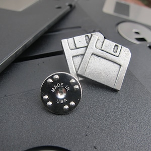 May include: Silver-colored floppy disk pin and backing, with the words "MADE IN USA" on the backing. The pin is a retro design, with two floppy disk shapes. The pin is on a black surface with other floppy disks.