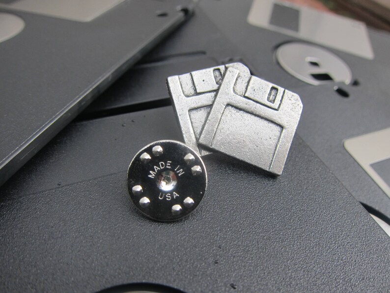 May include: Silver-colored floppy disk pin and backing, with the words "MADE IN USA" on the backing. The pin is a retro design, with two floppy disk shapes. The pin is on a black surface with other floppy disks.