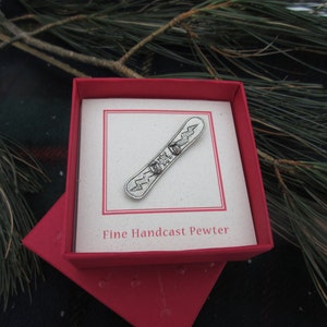 Puede incluir: Un amuleto de snowboard de peltre plateado en una caja roja con las palabras "Fine Handcast Pewter".