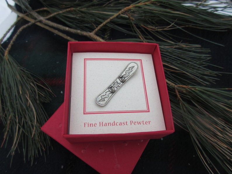 Puede incluir: Un amuleto de snowboard de peltre plateado en una caja roja con las palabras "Fine Handcast Pewter".