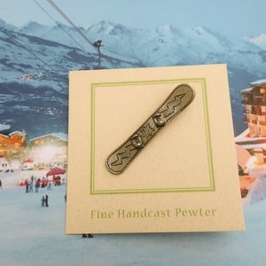 Puede incluir: Una figurita de esta&ntilde;o de una tabla de snowboard con un dise&ntilde;o de rayo. La figurita est&aacute; en una tarjeta que dice "Fine Handcast Pewter".