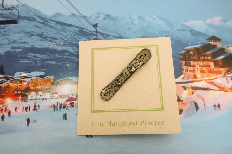 Puede incluir: Una figurita de esta&ntilde;o de una tabla de snowboard con un dise&ntilde;o de rayo. La figurita est&aacute; en una tarjeta que dice "Fine Handcast Pewter".