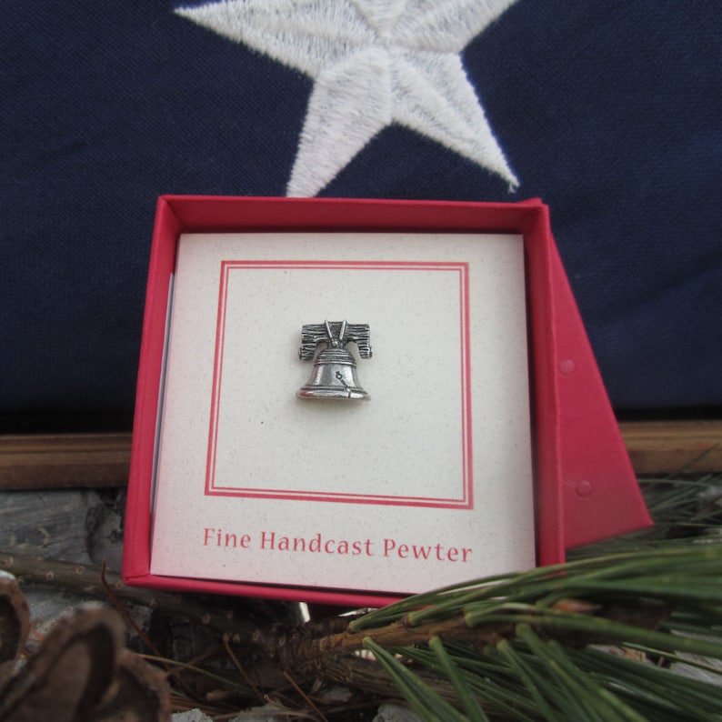 Puede incluir: Un peque&ntilde;o pin de plata de la Campana de la Libertad en una caja roja con fondo blanco. La caja tiene el texto "Fine Handcast Pewter" impreso en la parte inferior.