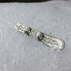 Puede incluir: Un amuleto de metal plateado en forma de tabla de snowboard con un dise&ntilde;o de rayo y estrella.