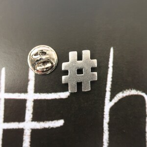 May include: Silver metal hashtag pin with a silver metal back. The pin is on a black background with a white hashtag drawn on it.