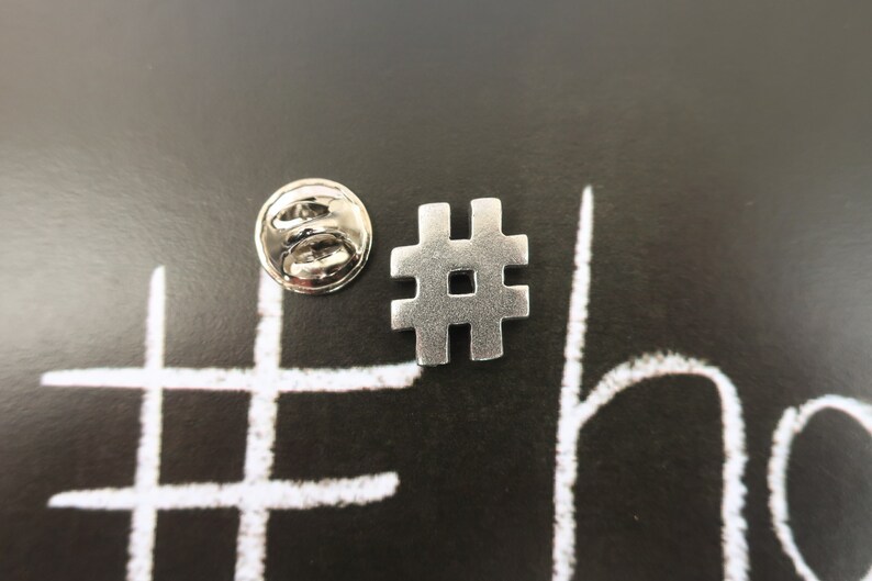 May include: Silver metal hashtag pin with a silver metal back. The pin is on a black background with a white hashtag drawn on it.