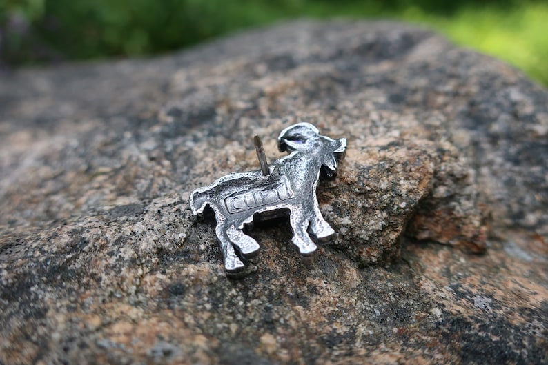 May include: A silver metal goat pin with a detailed design. The pin has a small metal pin on the back for attaching to clothing or a bag.