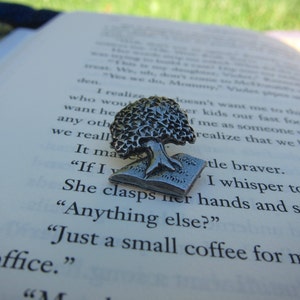 May include: A silver-colored tree pin, with detailed foliage, is set on top of an open book. The pin's design features a tree atop an open book. The book's pages display printed text. The pin is a decorative accessory.