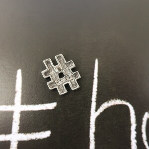 May include: A silver metal charm in the shape of a hashtag symbol. The charm is on a black background with a white hashtag drawn on it.