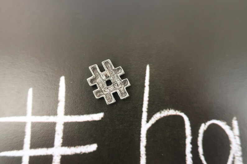 May include: A silver metal charm in the shape of a hashtag symbol. The charm is on a black background with a white hashtag drawn on it.