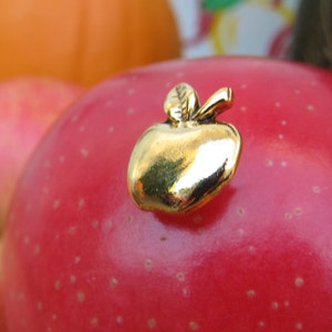 Puede incluir: Un pin de manzana de color dorado sobre una manzana roja. El pin de manzana tiene una hoja y un tallo.