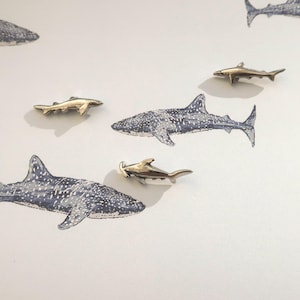 May include: A collection of small, gold-colored shark pins arranged on a white background with watercolor illustrations of whale sharks. The pins are detailed and shiny, contrasting with the softer, blue-grey painted sharks.