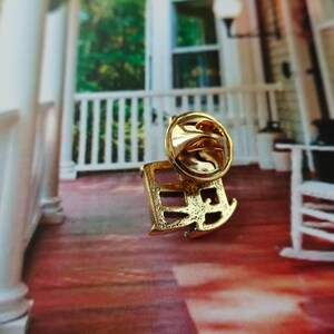 Pode incluir: Pin de metal dourado com um design estilizado de uma cadeira de balan&ccedil;o. O pin tem um fecho de seguran&ccedil;a na parte de tr&aacute;s.