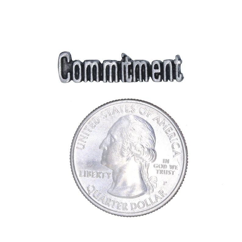May include: A silver charm with the word "Commitment" engraved on it. The charm is about the size of a US quarter.