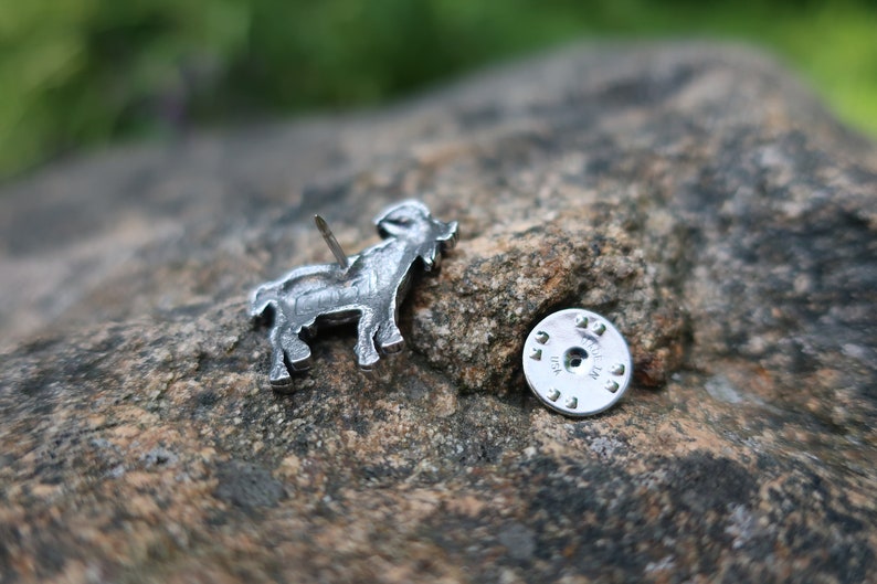 May include: A silver metal goat pin with a detailed design. The pin has a back with a silver metal clasp and a pin.