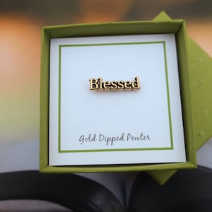 Puede incluir: Un pin de peltre bañado en oro con la palabra "Blessed" en letras en relieve. El pin se muestra en una caja verde con una tarjeta blanca que dice "Gold Dipped Pewter".