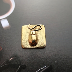 May include: A gold-colored metal pin shaped like a computer mouse on a square pad. The mouse has a cord that is curled up.